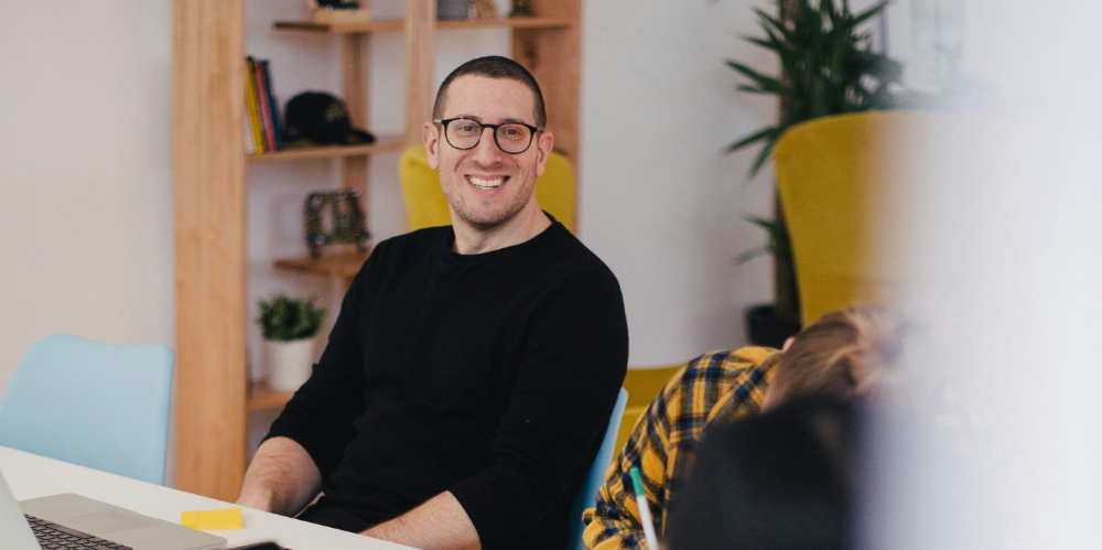 Man in black top and glasses, sitting at a table, relaxed and smiling to camera  