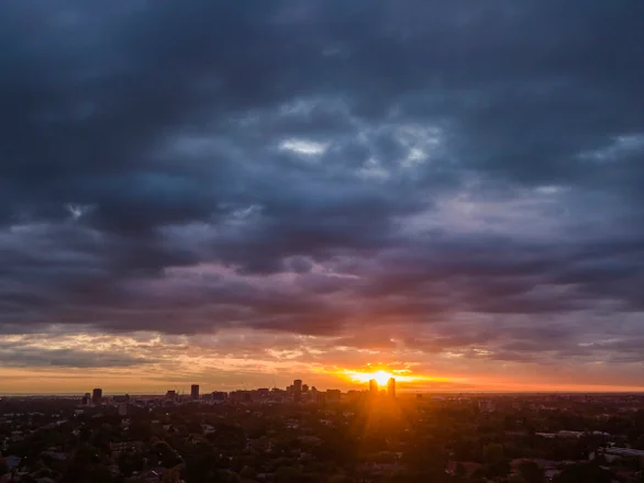 The autumn/summer sun sets beneath dark, moody skies over the Adelaide Central Business District seen in the distance from the leafy eastern suburbs.