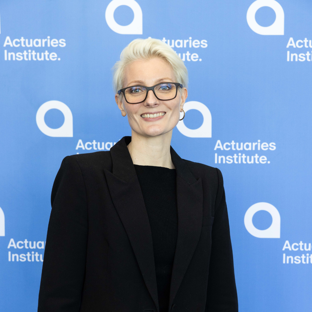 Female with short blonde hair, glasses smiling at camera in from of a company banner