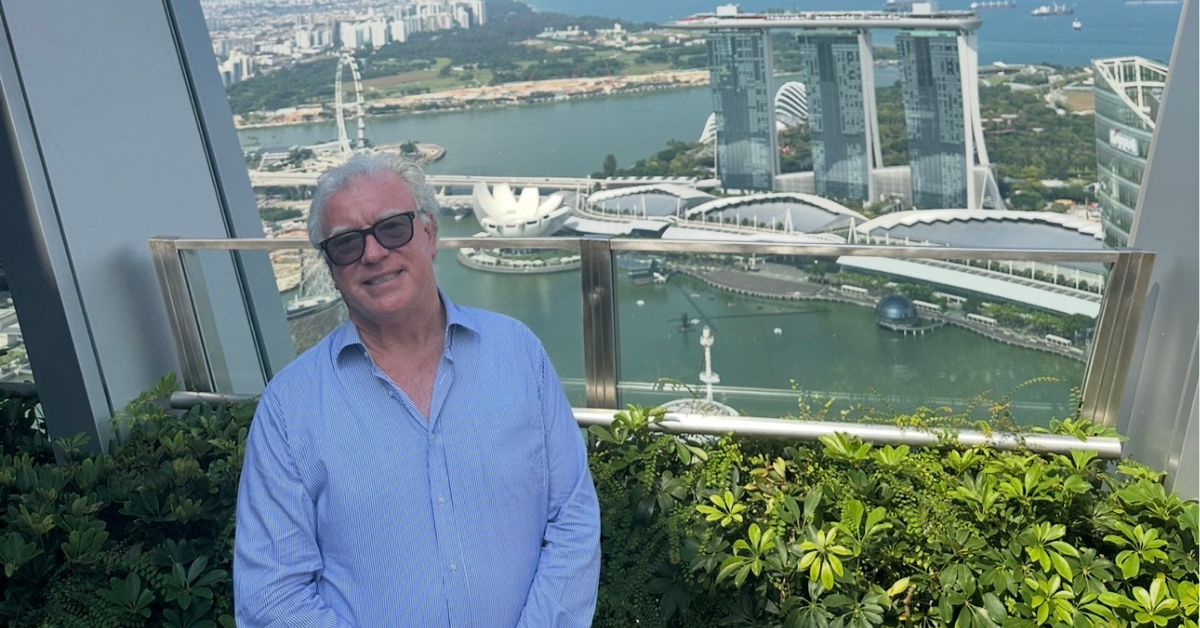 John Maroney standing on a high-rise terrace with the Singapore skyline, Marina Bay Sands, and the Singapore Flyer visible in the background.
