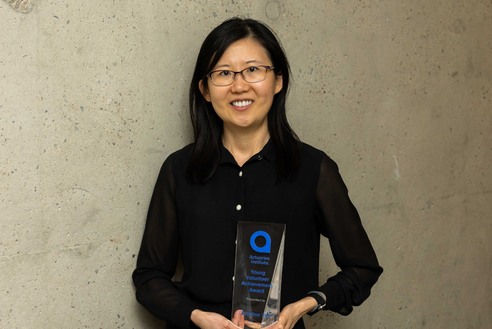 Female with black dress, hair and glasses, smiling at camera and holding a trophy