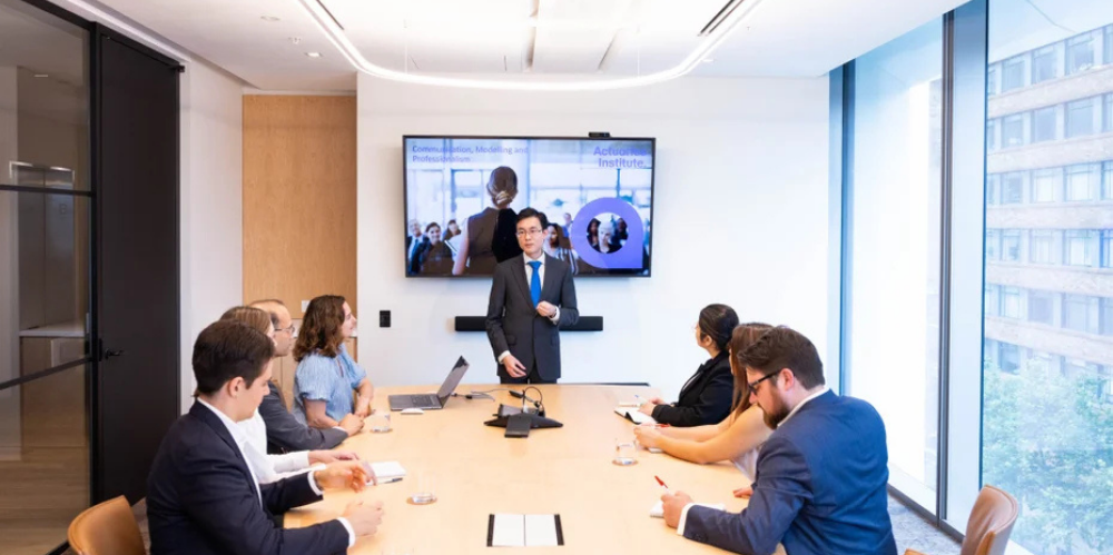 A man presents to a group of six colleagues seated around a boardroom table, with an Actuaries Institute slide displayed on a screen behind him.
