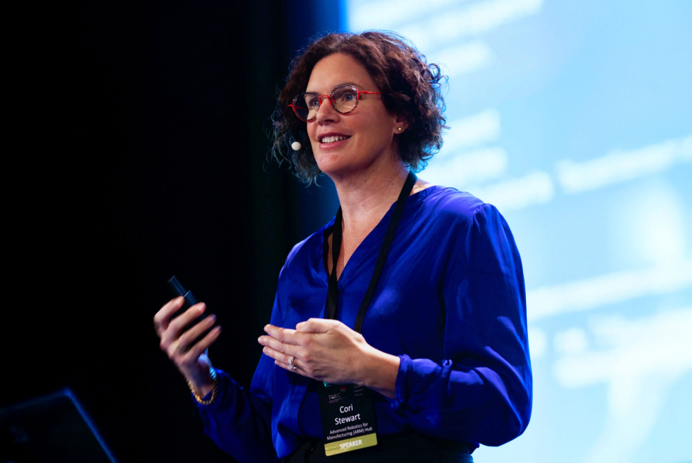 Female with short brown curly hair on stage speaking to audience in front of a screen, dresses in cobalt blue shirt