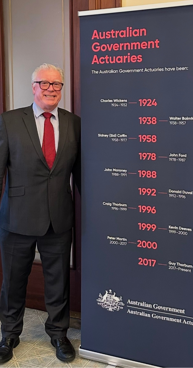 John Maroney standing beside a display banner listing all Australian Government Actuaries since 1924, with his name listed for the period 1988–1991.