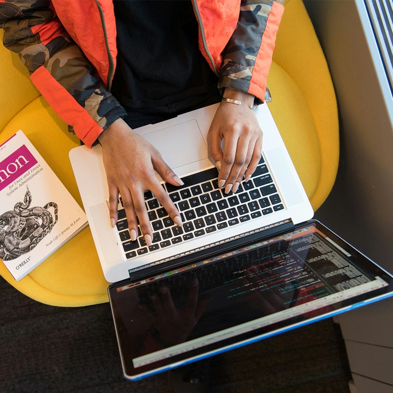 Someone typing on laptop coding with python book next to them