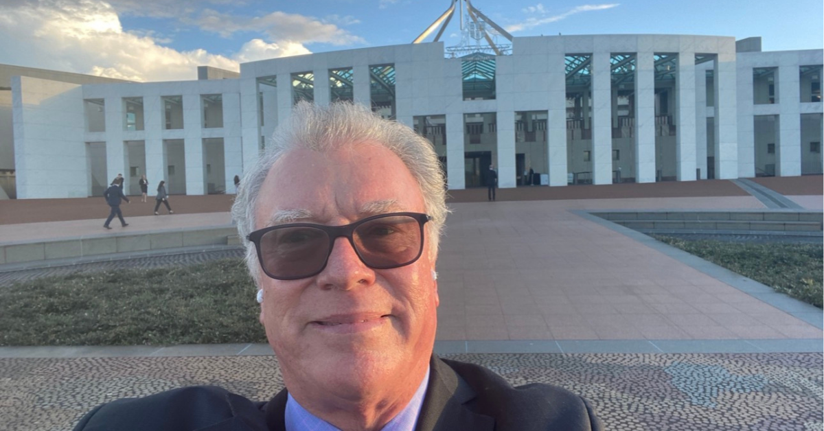John taking a selfie outside Parliament House in Canberra on a partly cloudy day, with the building's flagpole visible in the background.