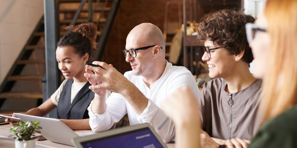 Four people sitting at a desk all looking off to the left with papers and computers on the desk