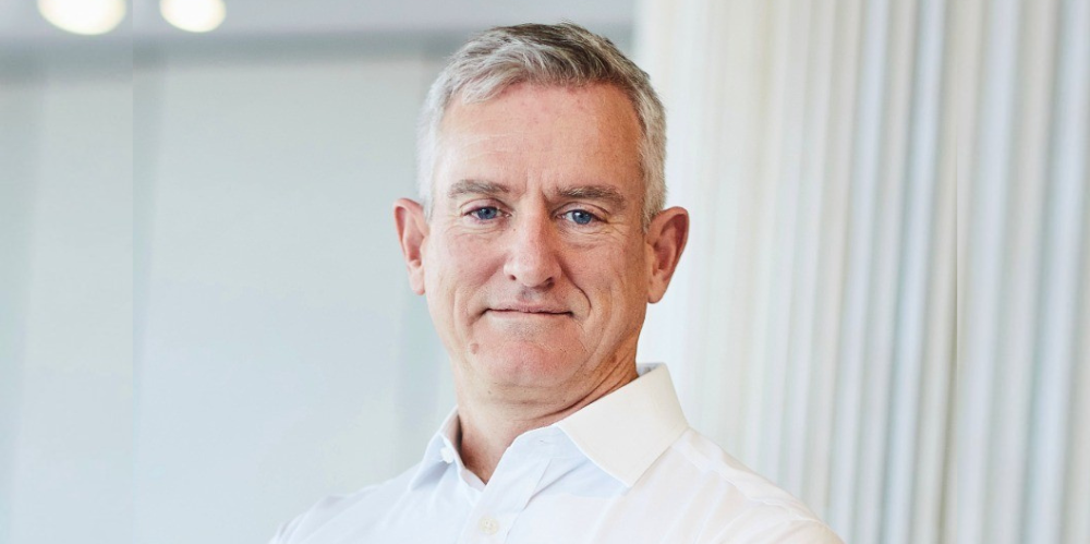 Male in white shirt smiling at camera with light coloured hair, and standing in front of white office background