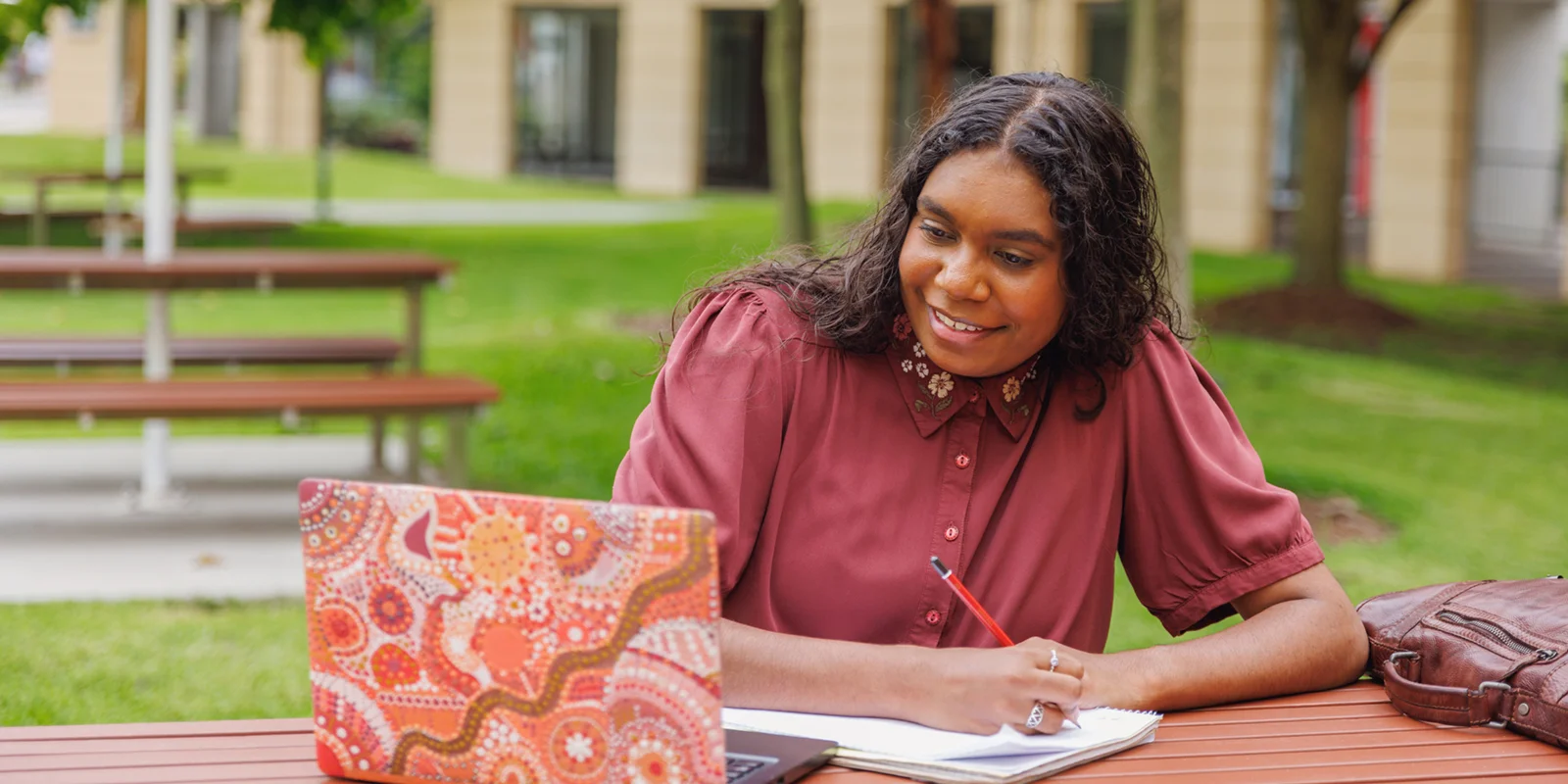 A smiling First Nations woman sits outside at a table writing in a notebook while looking at a laptop.