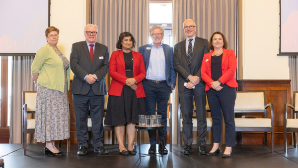 Standing on a conference stage, 6 speakers line up for a photo