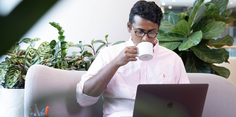 Man sitting on a couch, sipping from a mug while looking down at a laptop.