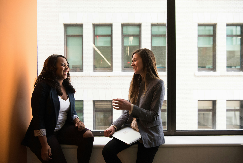 Two females sitting on a window bench chatting