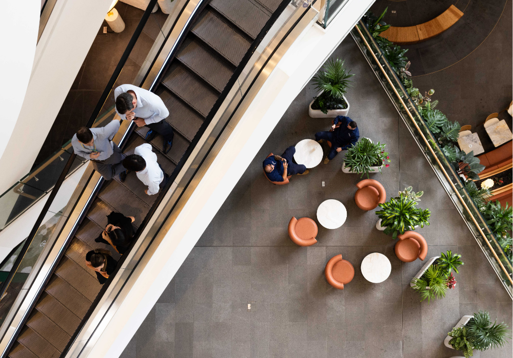 Overshot of a business foyer that captures people on an escalator and others having coffee. 