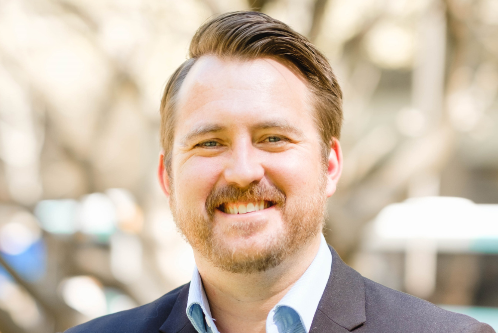 Photo of business man to shoulder in outside setting.He has light brown hair and beard, is smiling wearing a grey suit and light blue shirt.