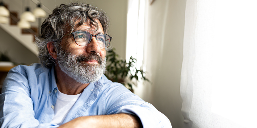 Thoughtful mature man with eyeglasses and gray hair looking outside the window while sitting at home.