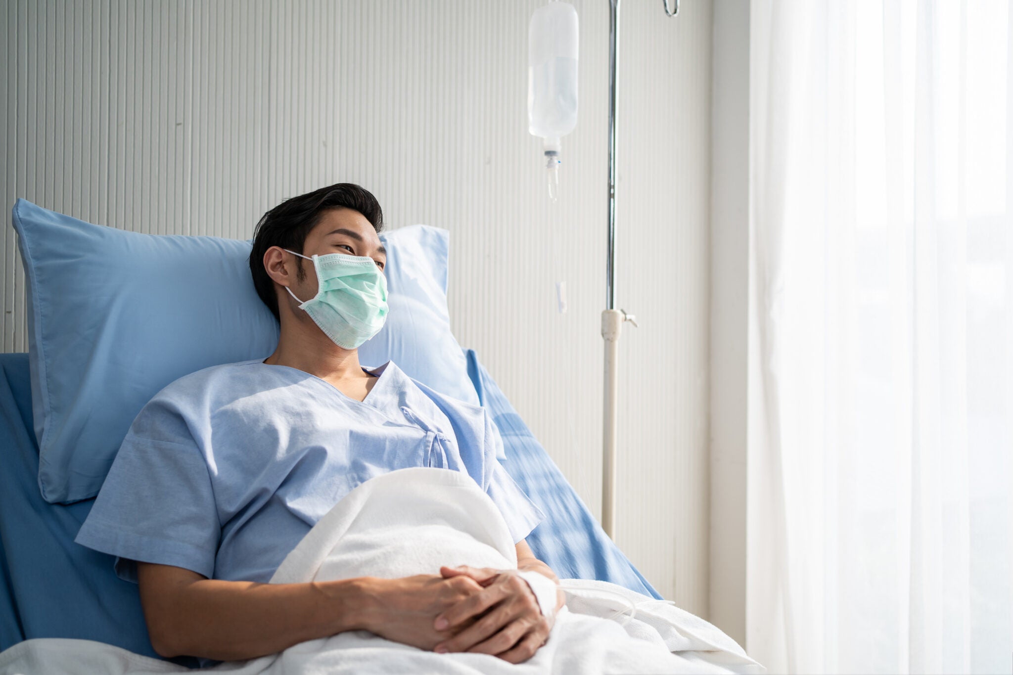 Asian young male patient wearing protective face mask, lying on bed in the hospital and getting saline solution in medical process. He has infected from the Coronavirus with feeling hope to get well.
