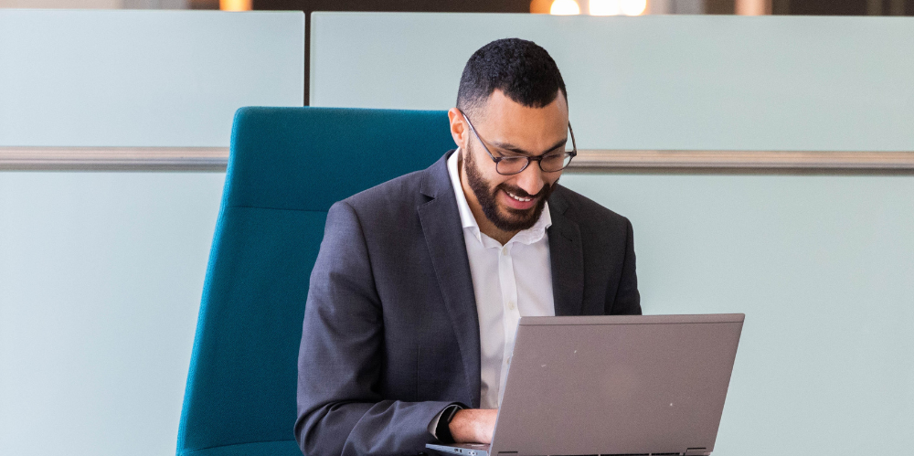Male with dark hair abd beard, in a suit looking down at his computer and smiling, working