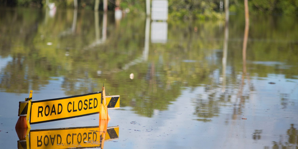 A flooded road with a road closed sign nearly being ingulfed by the flood waters.