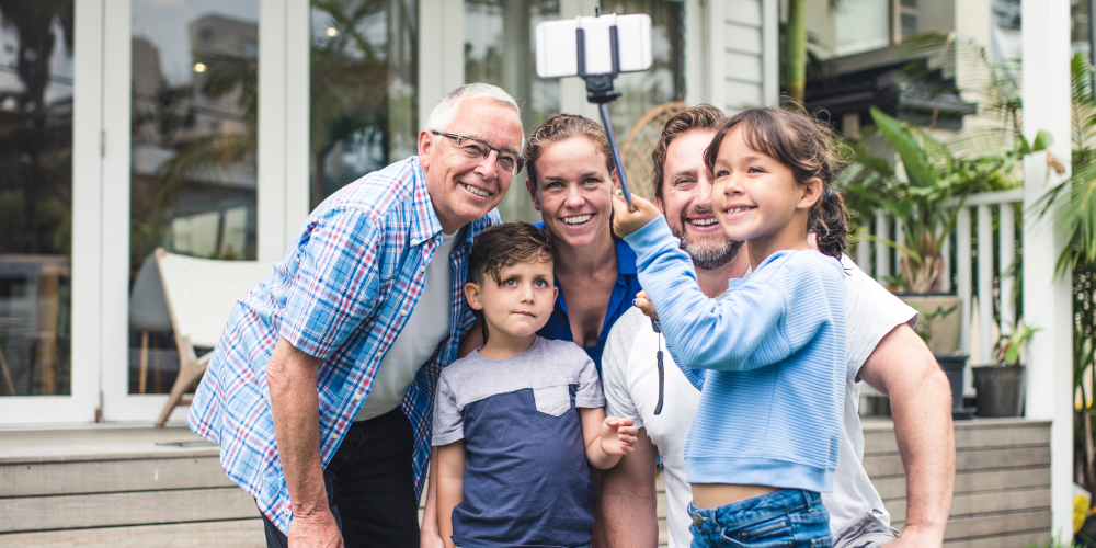 A three-generation family of five — a grandfather, two parents and two young children — pose together for a selfie on a stick outside a light-coloured weatherboard home. All are smiling and casually dressed. Lush green plants are visible in the background.