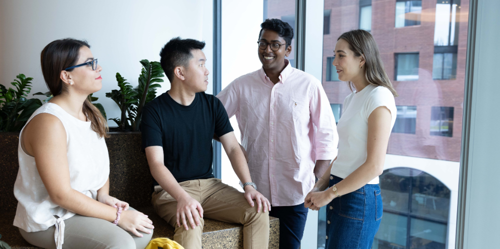 A group of young people seated and standing in a office area, casually dressed. From left is a woman, 2 men and a woman on the right.