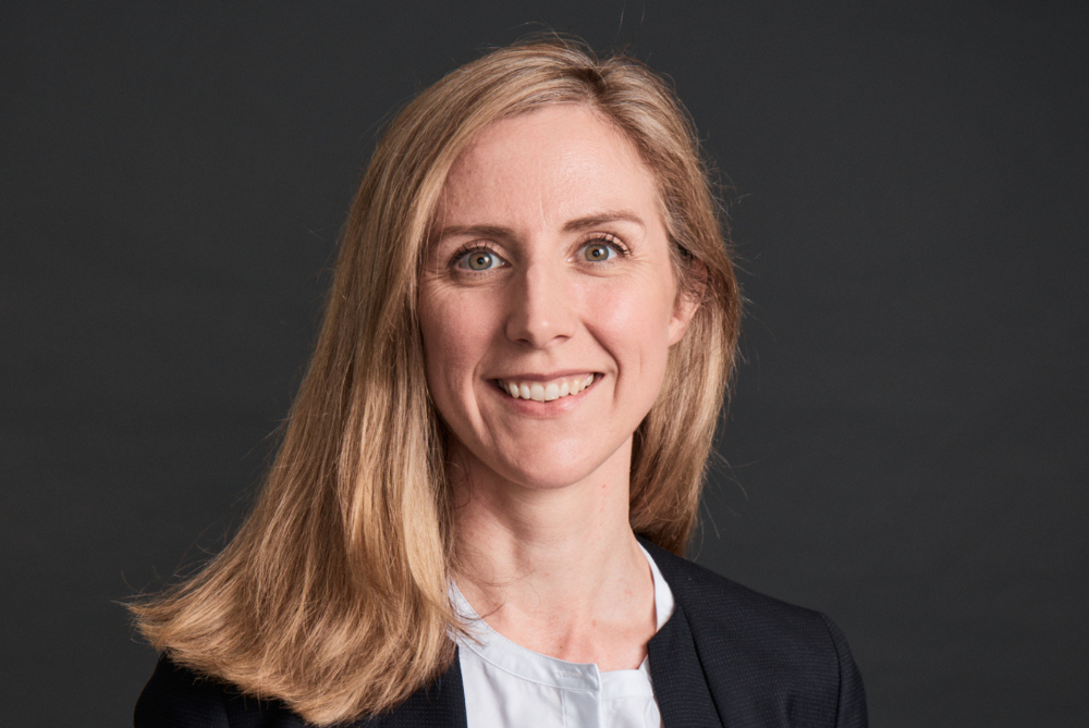 Women with blond hair to shoulders smiling and looking at camera. Dark grey background.