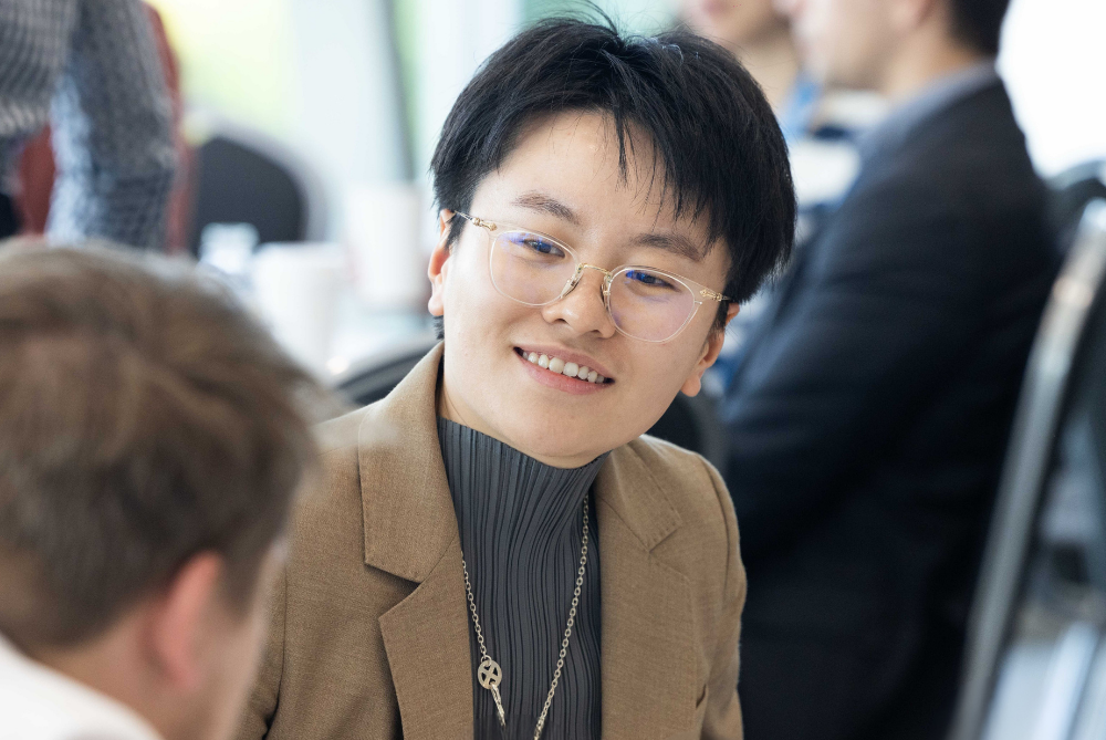 Female with short balck hair, glasses and brown jacket, sitting and leaning forward smiling