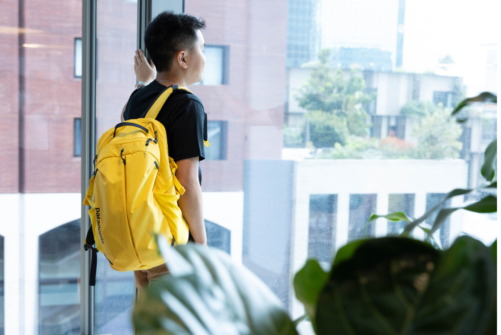 Young man in black tshirt looking out an office window with a yellow backpack over shoulder