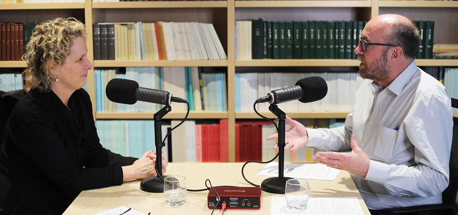 Margo Lydon and Geoff Atkins in conversation at a podcast recording desk with microphones, with Geoff gesturing as he speaks.