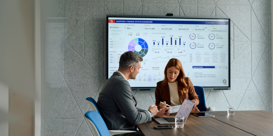 Two business colleagues engaged in a corporate meeting, analysing financial data presented on a large screen and their personal laptops, collaborating on business strategy