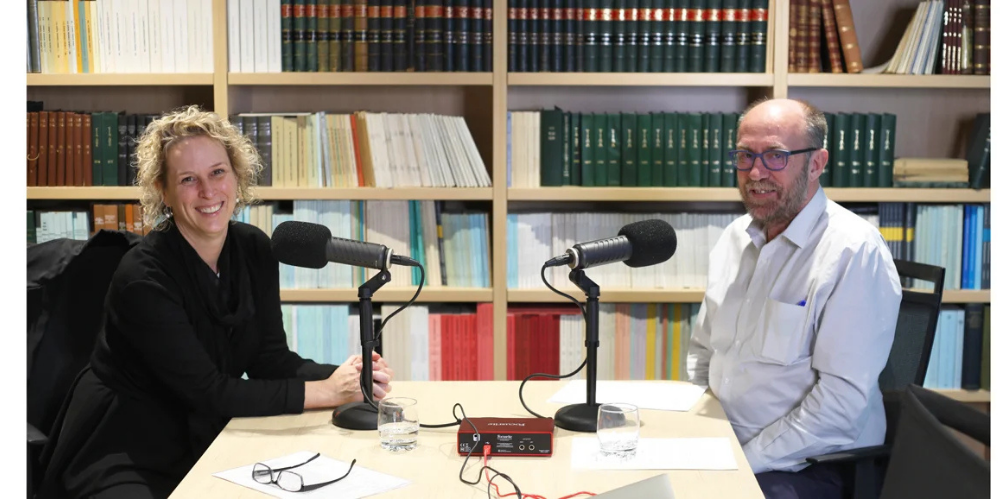 Margo Lydon and Geoff Atkins seated at a podcast recording desk with microphones in front of a bookshelf.