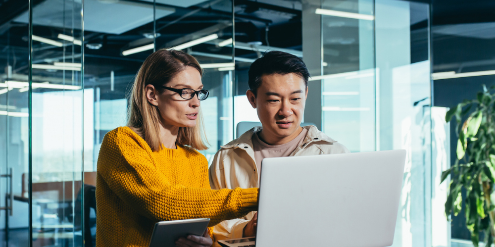 Two people in office looking at laptop, female blonde hair and black rimmed glasses with yellow jumper is talking and male with dark hair and cream casual jacket is looking at the screen.