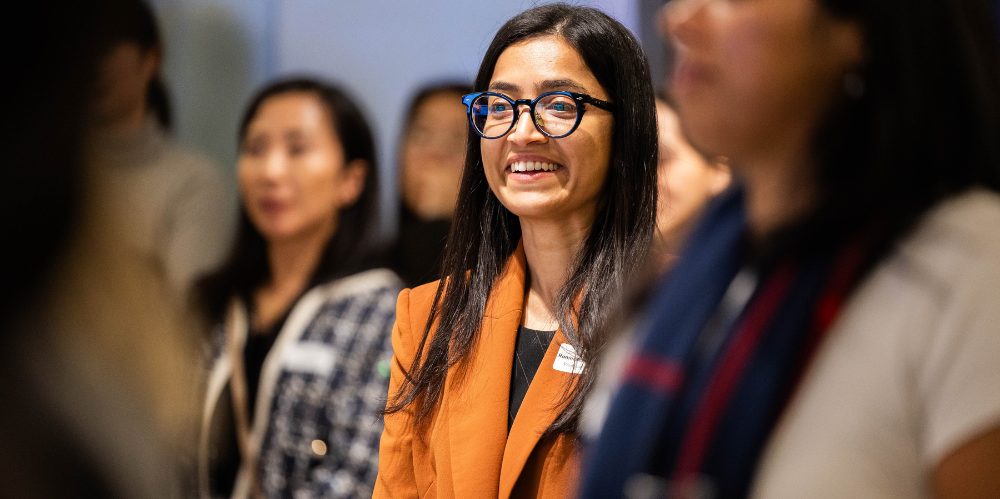 Business women with long black hair, black glasses and orange jacket standing at an event and smiling off camera