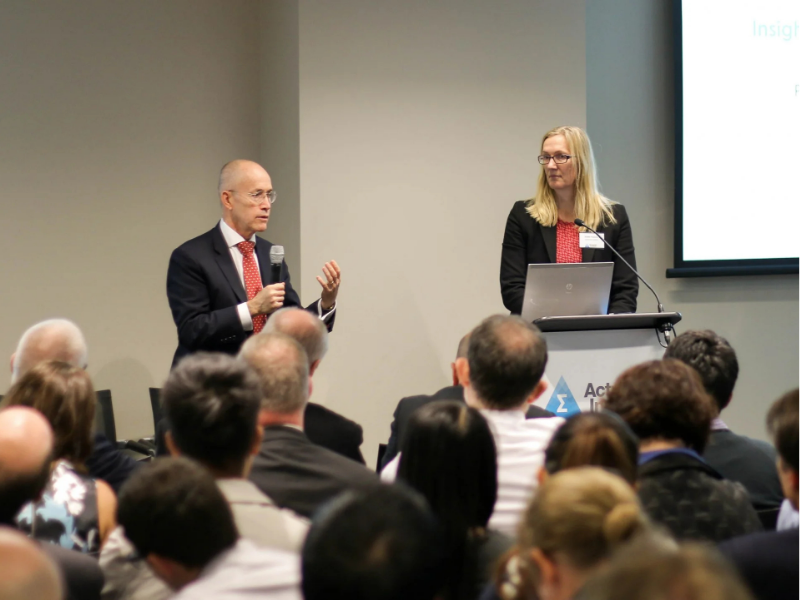 A man speaks into a microphone beside a woman presenting at a podium at an Actuaries Institute event, with a full audience in the foreground.