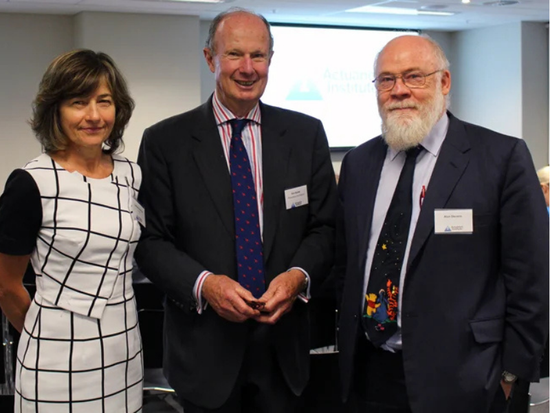 Three people — two men and a woman — smile for a photo at an Actuaries Institute event, with branded signage visible in the background.