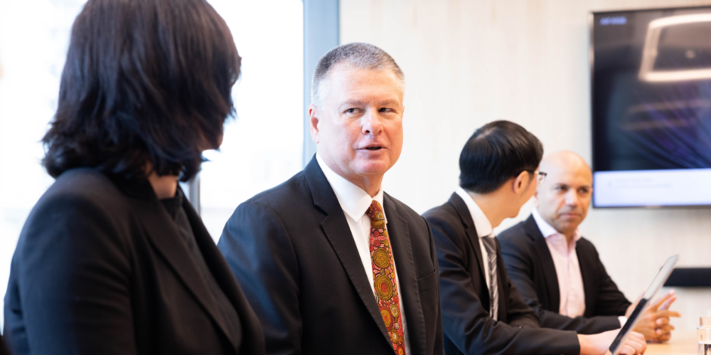 People sitting in board room, and one man with suit, white shirt and red and brown patterned tie looking to left and talking to business woman