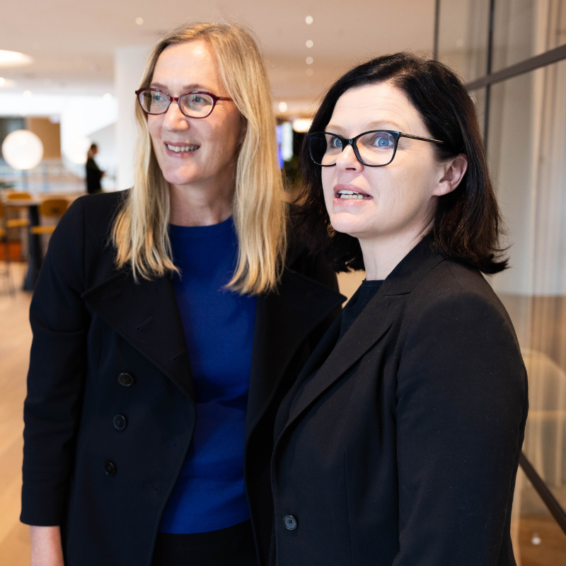 Two business women in office looking away and chatting, one with blonde hair and the other with dark hair