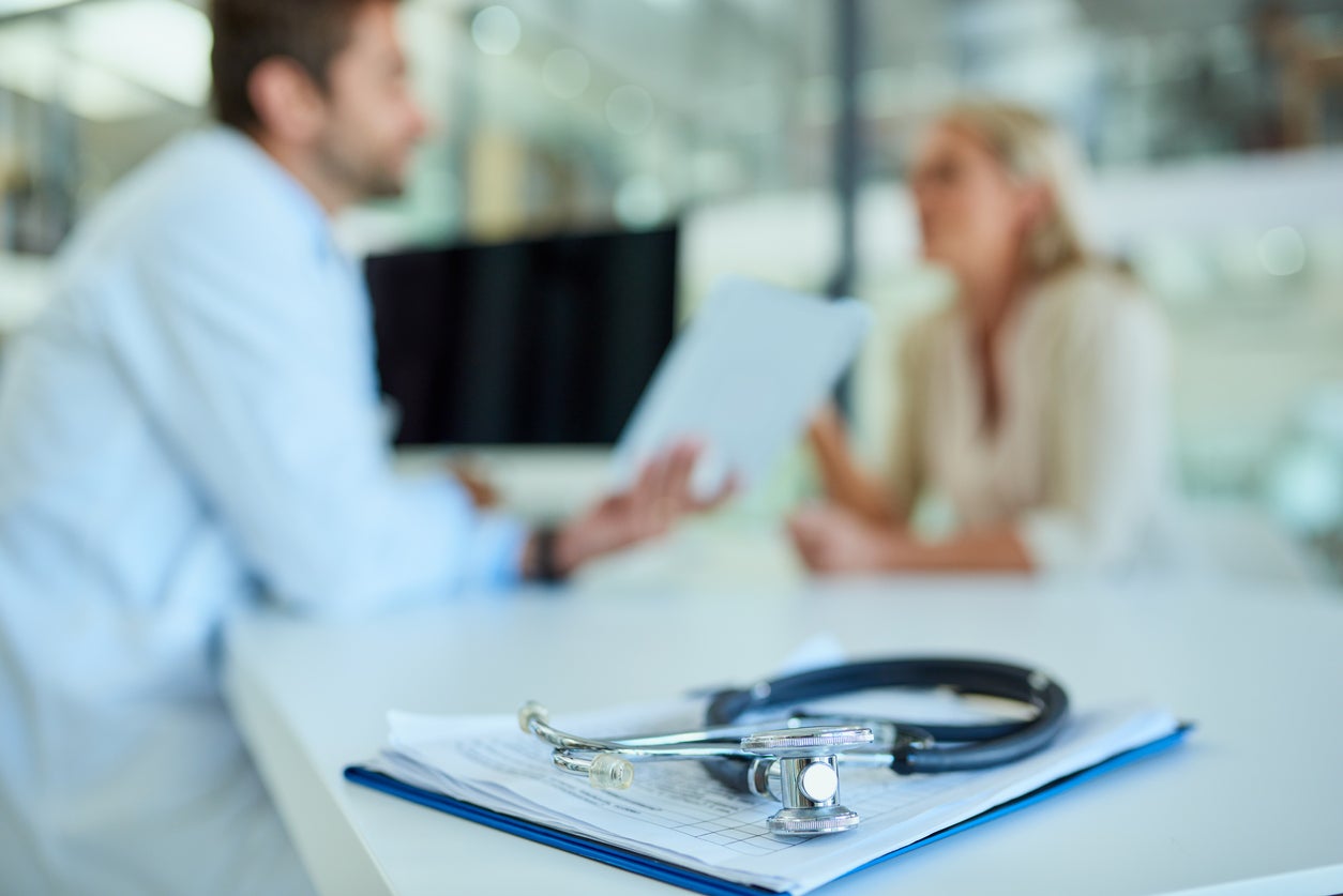 Shot of a stethoscope and a clipboard on a desk with a doctor and patient in the background