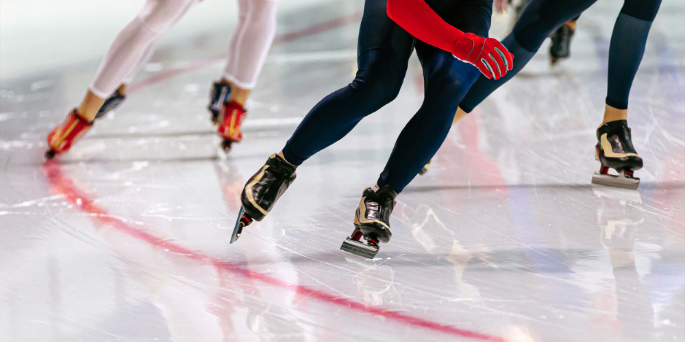 Close-up of speed skaters' legs and skates mid-race on an ice rink, showing powerful strides and reflections on the ice surface.