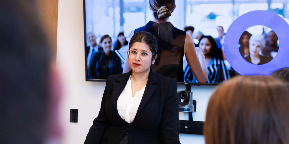 Female standing at a table with hands on table in black suit and white shirt talking to people, with screen behind her