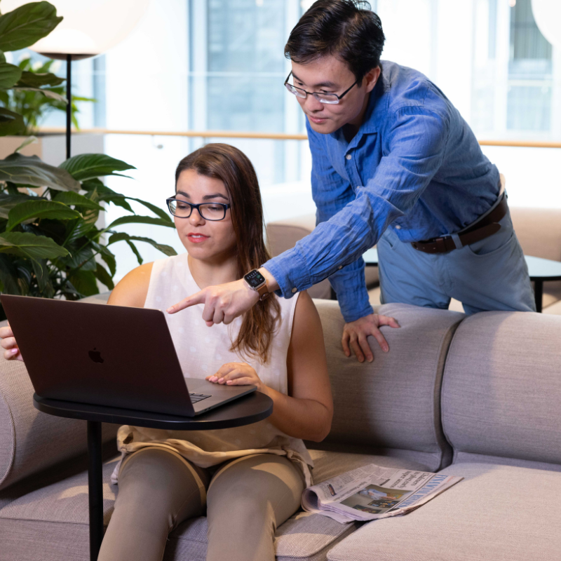 Business woman sitting on the lounge with a computer and a man in blue pants and shirt leaning over smiling and pointing to the computer
