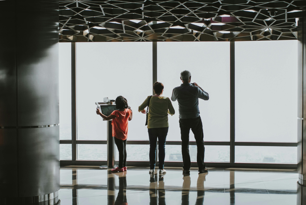 Image of two adults and a child looking at the view out of the window of a high rise building.