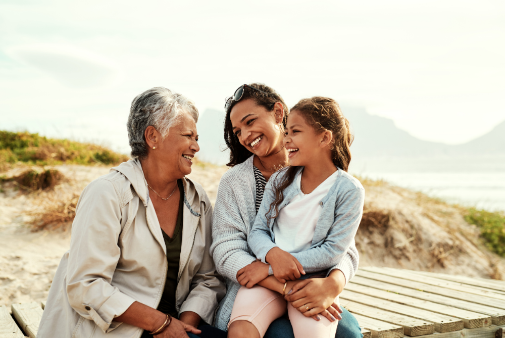 Three generations of women — a grandmother, mother and young girl — sit together on a beach boardwalk, smiling and laughing in warm afternoon light.