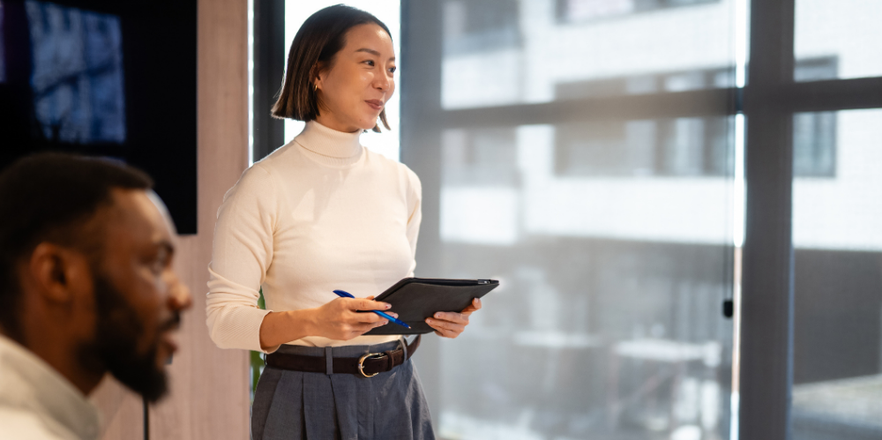 Asian business woman standing, holding a tablet and a pen, leading a presentation during a corporate team meeting in an office