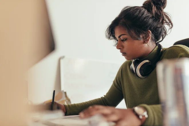 Woman making notes while working on computer. Female computer programmer working at her desk.