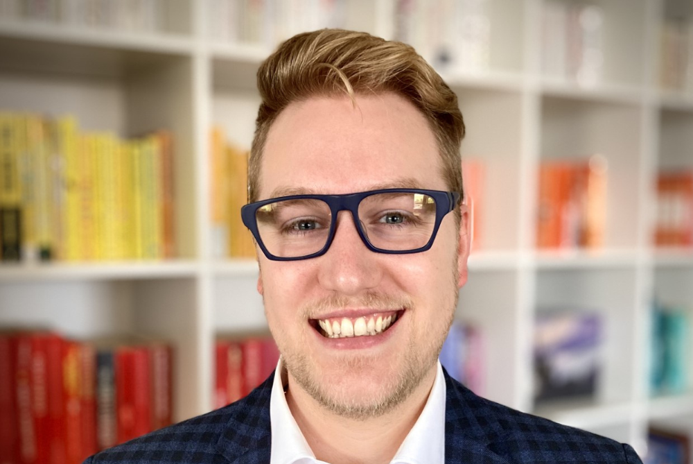 Male head shoulders photo, set with a coloured coded bookshelf in the background. Man is smiling with short brown hair, dark glasses amd wearing a checked jacket and white coloured shirt.