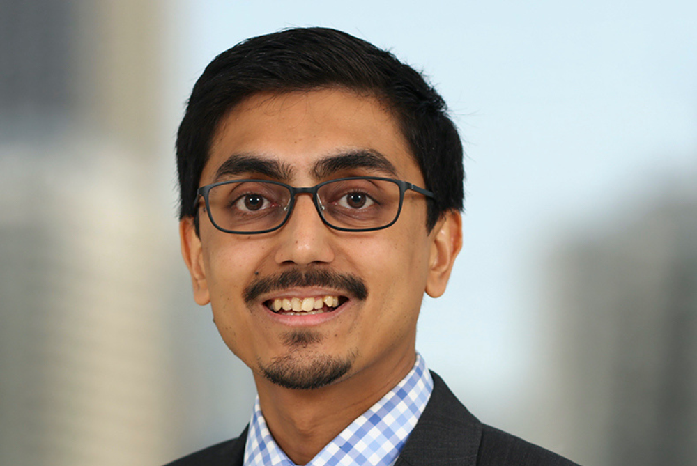 Portrait photo of male, smiling with black glasses, moustache and small beard, and checked shirt and jacket.