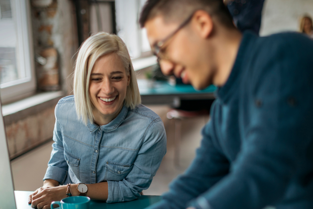 Women blond hair and denim shirt, leaning on desk with computer, smiling and talking to a male colleague who is sharing a document
