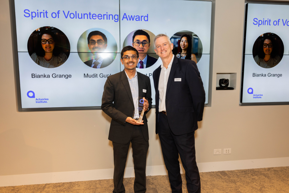Two business men in suits in front of a screen, smiling and one holding a trophy