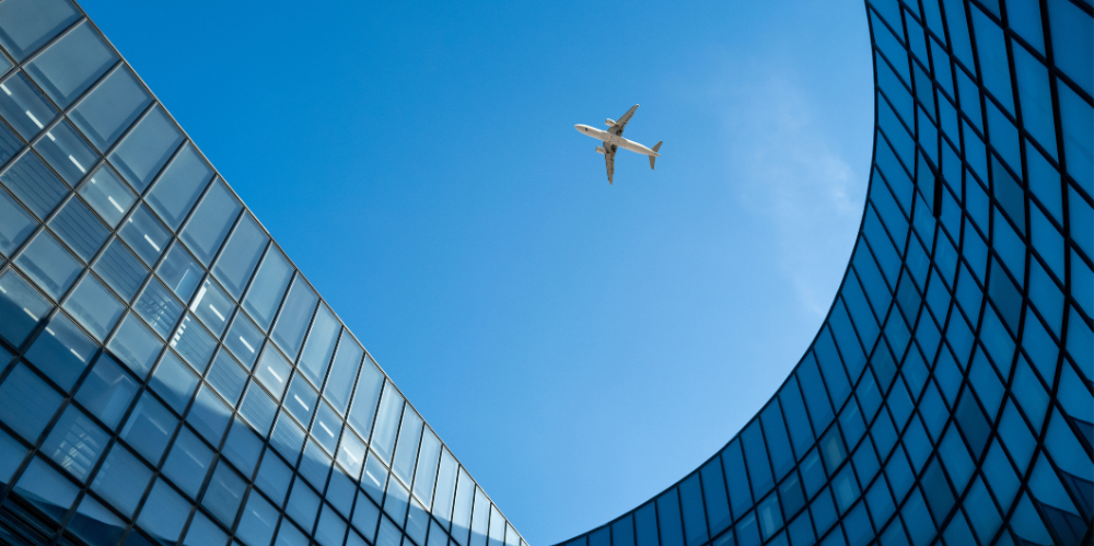Image depicts an airplane soaring through a clear sky above a corporate building