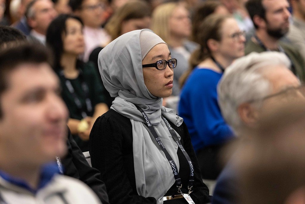 Photo of a seminar or session where hundreds of people are listening intently, the focus of the photo is on a woman in the sea of listeners